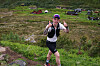 En løper med sekk og caps beveger seg på en fjellsti med grønt landskap og biler i bakgrunnen.
