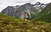 En løper i rødt og sort løper på en gresskledd fjellside med høye, snødekte fjell bak.