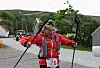 En person i rød jakke og sekk løper med staver på en vei ved grønnkledde fjell.
