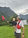 En mann holder et lite barn som vifter med et norsk flagg foran fjell og fjord.