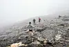 Flere personer beveger seg over et steinete fjell i tett tåke.