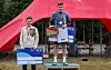 Tre unge deltakere med medaljer står på en pall under en rød teltduk.